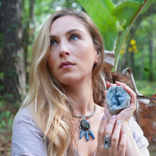 Beautiful woman poses with a crystal geode in her hand while wearing a pendant and ring designed by a woman-owned jewelry designer and wholesaler. 